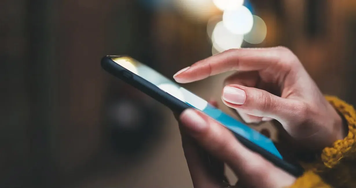 A close-up of a person’s hands using a smartphone, with one hand holding the phone and the other hand touching the screen. The background is blurred with bokeh lights.