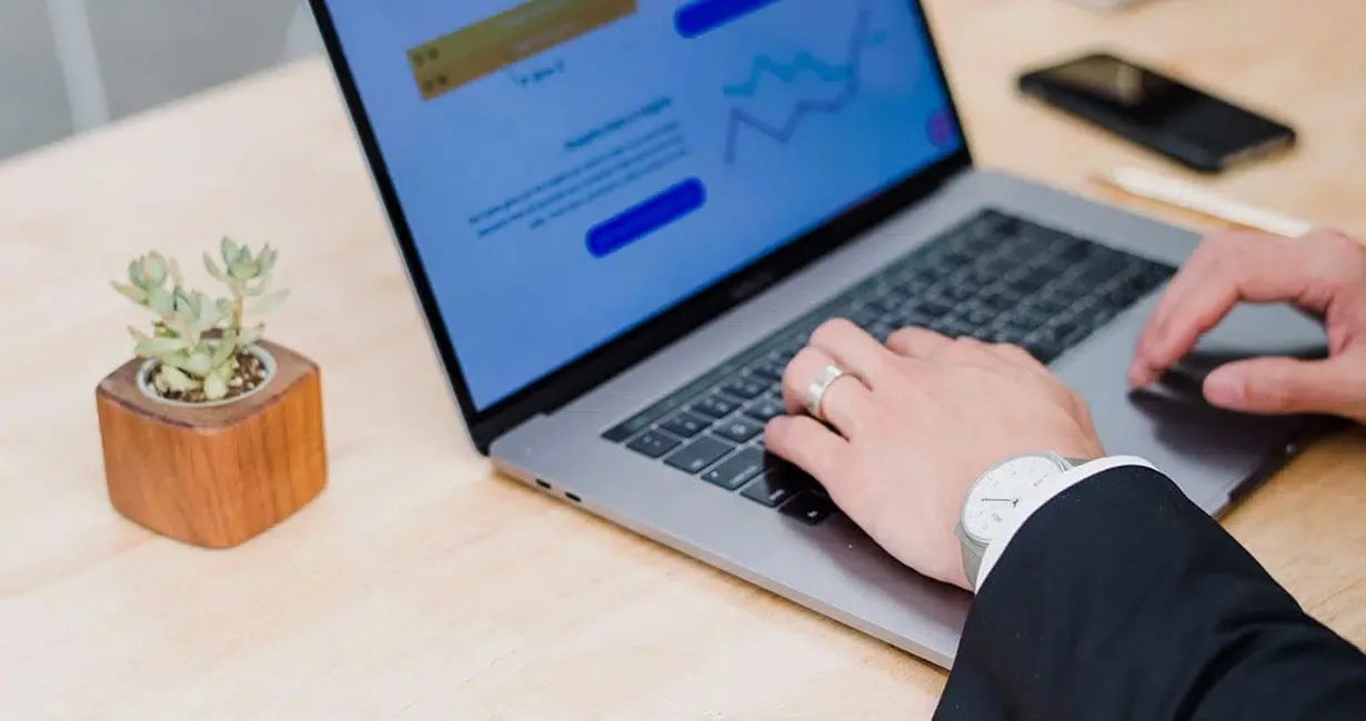 A person in a suit types on a laptop at a wooden desk. A small potted plant, a smartphone, and a pen are nearby. Graphs and charts are visible on the laptop screen.